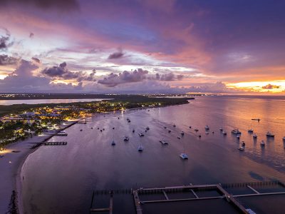 Aerial view of a coastal marina at sunset with boats anchored in calm waters, beachfront resort lighting, and a dramatic purple and orange sky